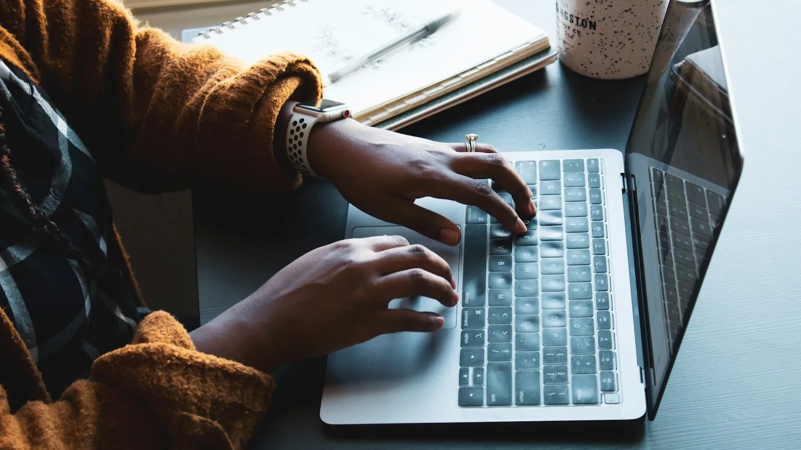 A person types on a laptop next to a notebook and a mug of coffee.